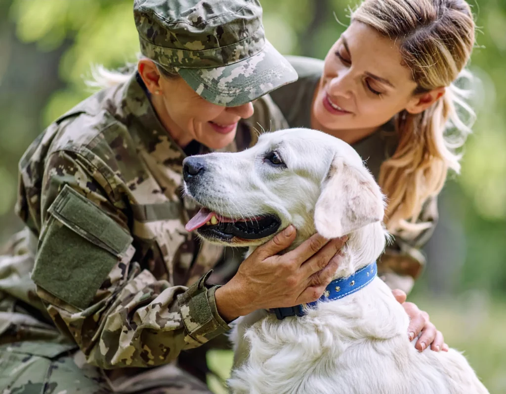 Accueil 3 Soldate blessée souriant en recevant un chien d’assistance, accompagnée d’une camarade en tenue militaire, dans un moment de soutien et de résilience.